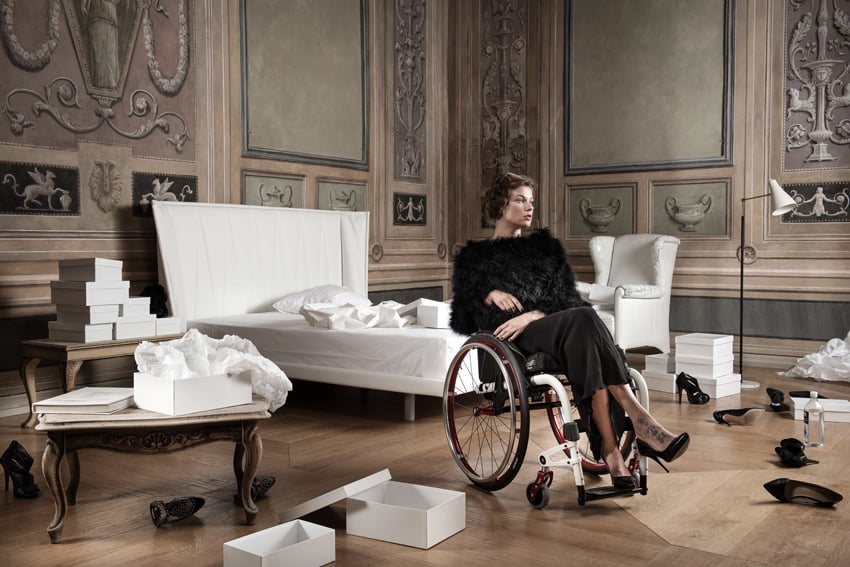 An image of a formally dressed woman wearing a black gown, black fur stole, and black pumps sitting with legs crossed in a wheelchair. She sits in a bedroom with a high ceiling with many open shoe boxes and pairs of heels scattered about.
